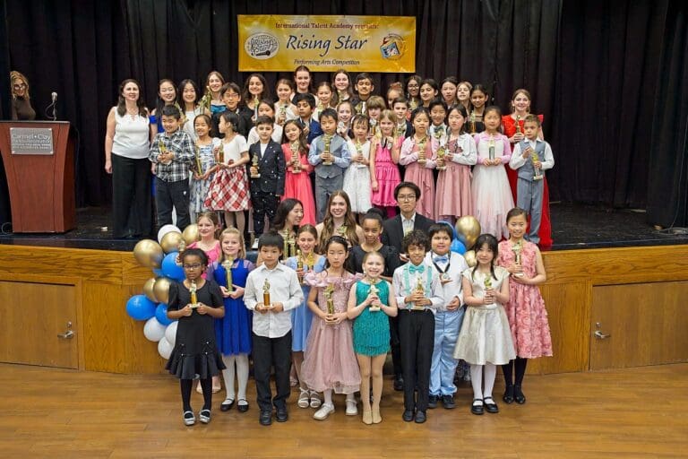 Group of children on a stage holding trophies posing for a photo after a performance with a yellow banner in the backdrop and balloons nearby